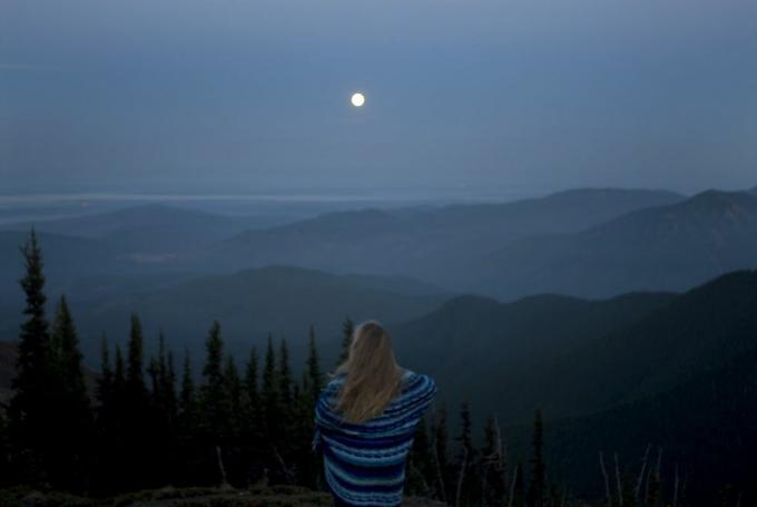 אישה עטופה בשמיכה מביטה בנוף הררי עם ירח מלא, מבט אחורי