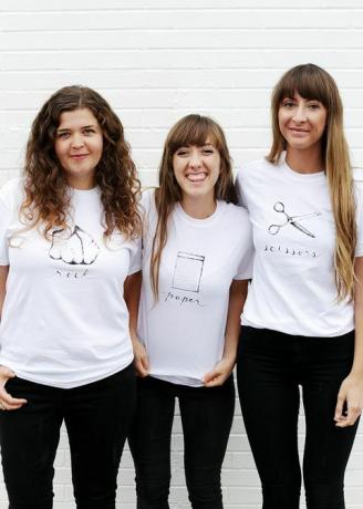 שלוש נשים בחולצות לבנות, עם סלע, ​​נייר או מספריים שנכתבו ותמונות על החולצה