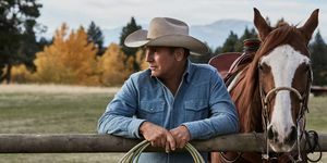 קווין קוסטנר באבן ילוסטון ליד סוס נשען על גדר עם חבל בידיים לבוש חולצת ג'ינס כחולה דהויה וכובע בוקרים בז'