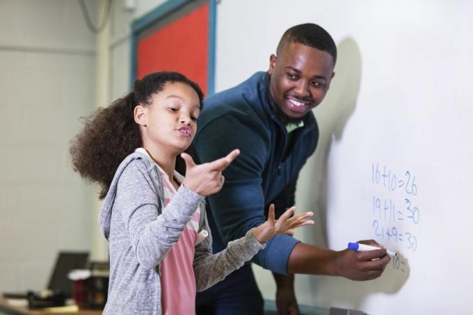 ילדה רב-גזעית בת 9 בשיעור מתמטיקה, עומדת ליד הלוח הלבן עם המורה שלה, גבר אפרו-אמריקאי צעיר בשלו בשנות ה-20 המורה מסתכל על תלמידו היסודי, מחכה בזמן שהיא מנסה להבין את התשובה, הפוקוס הוא על ילדה